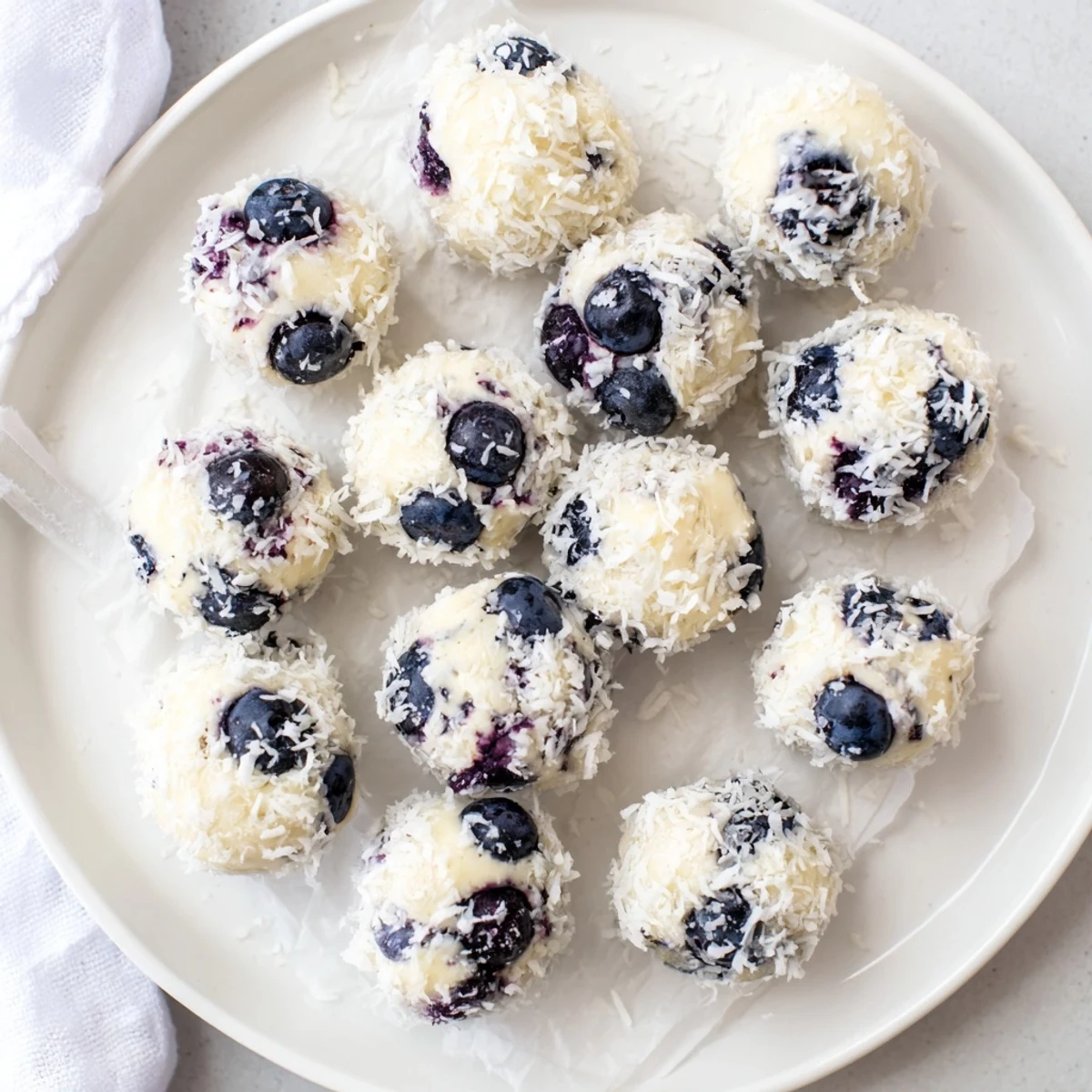 Arrangement of chilled blueberry cheesecake protein bites on white parchment paper topped with lemon zest and rolled coconut flakes
