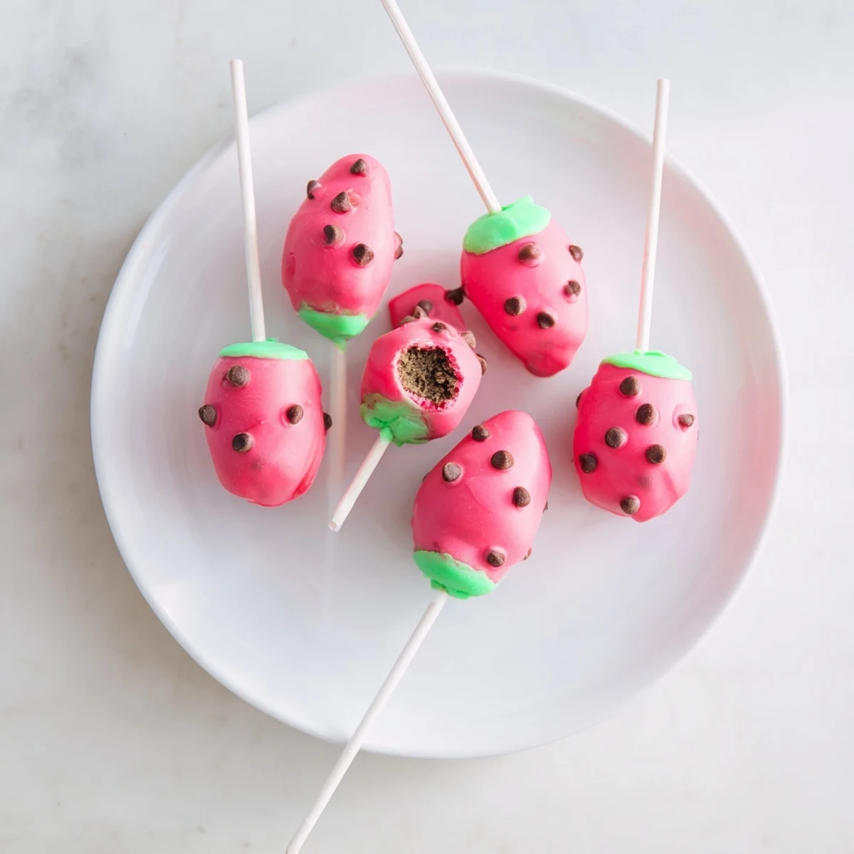 Pink watermelon cake pops decorated with chocolate seeds on green candy rind displayed on white platter