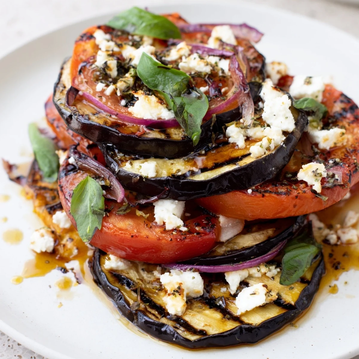 Smoky grilled tomato eggplant stacked on a platter with crumbled feta and fresh basil leaves