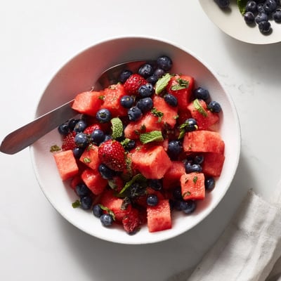 Summer watermelon fruit salad with berries, fresh mint leaves, and citrus honey dressing on white plate